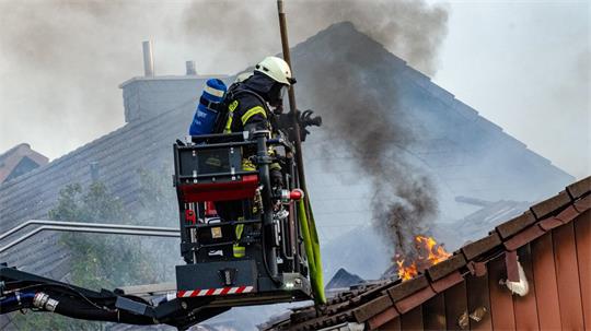 Feuerwehrmann in Schutzkleidung auf Hubsteiger löscht Dachbrand mit Flammen und Rauch.