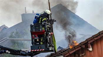 Feuerwehrmann in Schutzkleidung auf Hubsteiger löscht Dachbrand mit Flammen und Rauch.