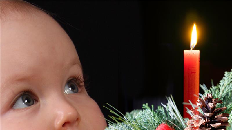 Ein kleines Kind schaut interessiert auf eine leuchtende Kerze auf einem Weihnachtskranz.