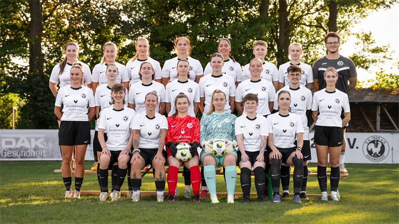 Gruppenfoto einer Frauenfußballmannschaft mit zwei Torfrauen in der vorderen Reihe auf einem Fußballfeld vor Bäumen