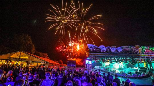 Menschenmenge bei Nacht vor einem beleuchteten Fahrgeschäft mit buntem Licht und Feuerwerk am Himmel