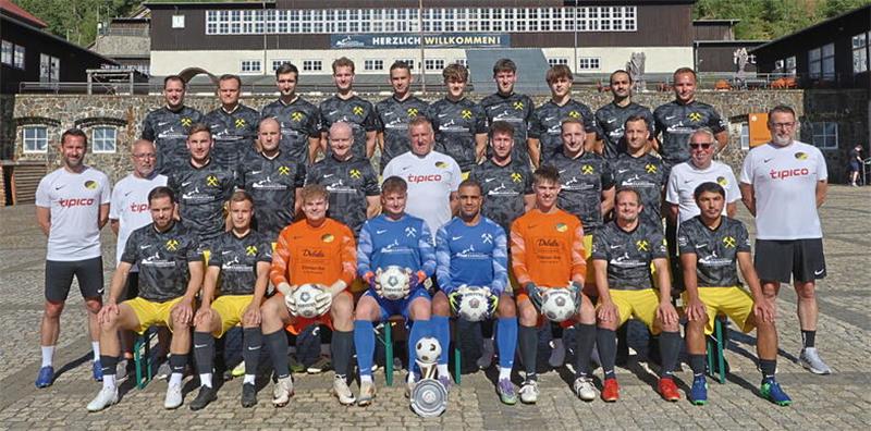 Gruppenfoto einer Fußballmannschaft mit Spielern in schwarzen Trikots und gelben Hosen sowie drei Torhütern in orangefarbenen und blauen Trikots vor einem historischen Gebäude mit Bergbauarchitektur
