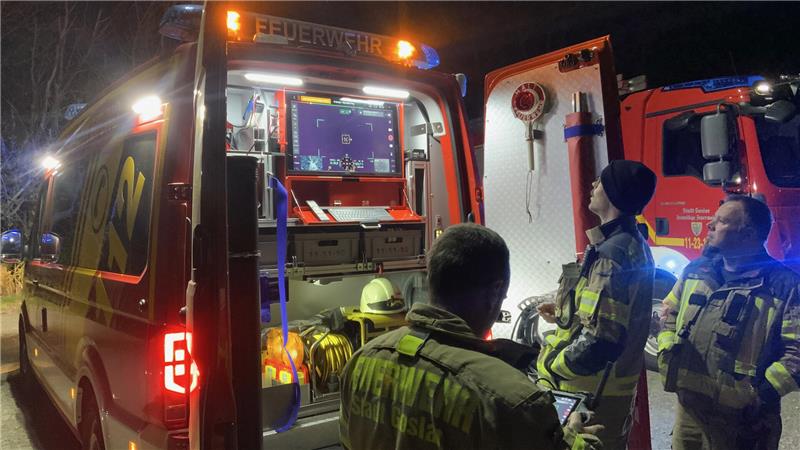 Drei Feuerwehrleute in Schutzkleidung stehen nachts vor einem geöffneten Einsatzfahrzeug mit beleuchtetem Innenraum.