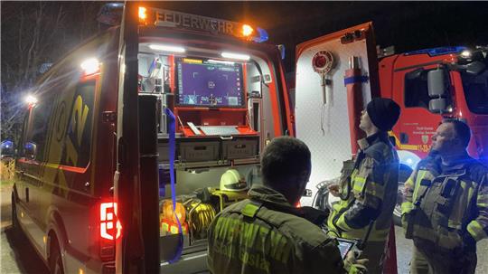 Drei Feuerwehrleute in Schutzkleidung stehen nachts vor einem geöffneten Einsatzfahrzeug mit beleuchtetem Innenraum.