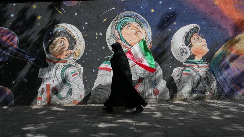 Mit einer iranischen Flagge in der Hand geht eine Frau an einer Wand vorbei, auf der Kinder in Raumanzügen zu sehen sind.