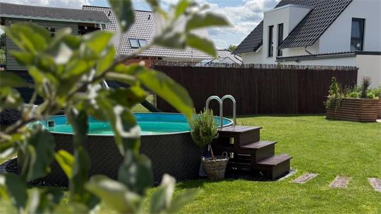 Garten mit grünem Rasen, aufgestelltem Pool mit Leiter und Treppe, im Vordergrund unscharfe Blätter, im Hintergrund weiße Häuser unter blauem Himmel mit Wolken.