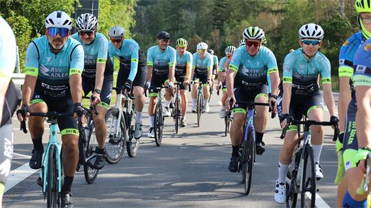 Gruppe von Radfahrern in hellblauen Trikots und Helmen fährt auf einer Straße in einer ländlichen Umgebung