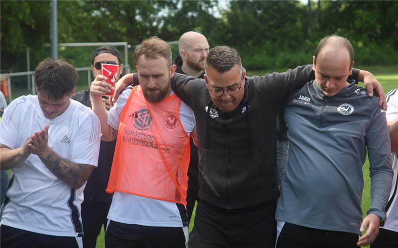 Gruppe von fünf Männern in Sportkleidung steht eng beieinander im Freien, einige klatschen und einer hält einen roten Becher
