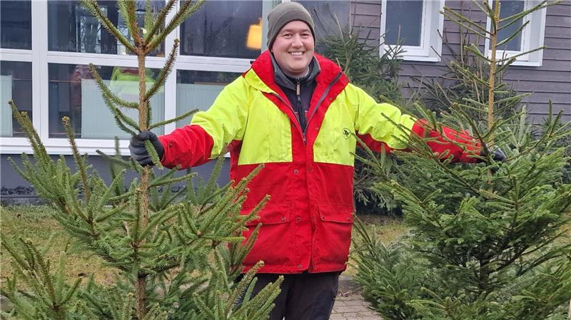 Ein Mann in einer gelb-roten Jacke hält grinsend zwei Bäume in der Hand.
