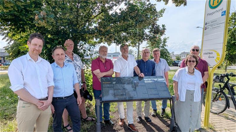 Gruppe von neun Personen steht im Freien neben einem Informationsschild unter einem Baum
