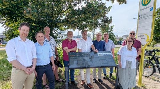 Gruppe von neun Personen steht im Freien neben einem Informationsschild unter einem Baum