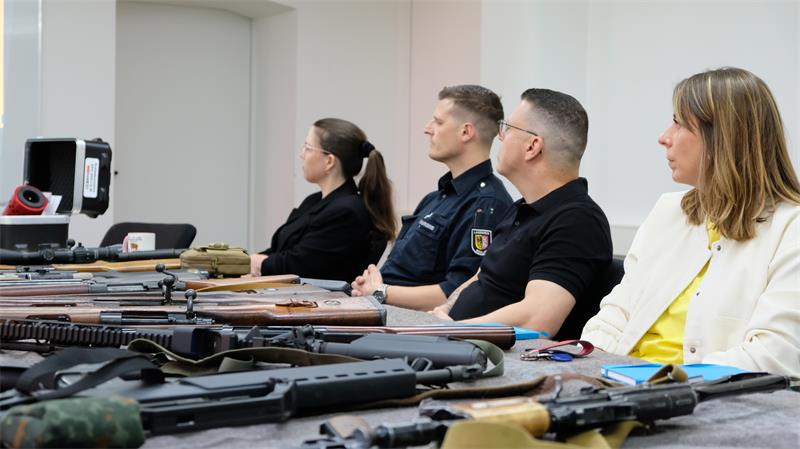 Zwei Frauen und zwei Männer sitzen an einem Tisch, vor ihnen liegen Gewehre.