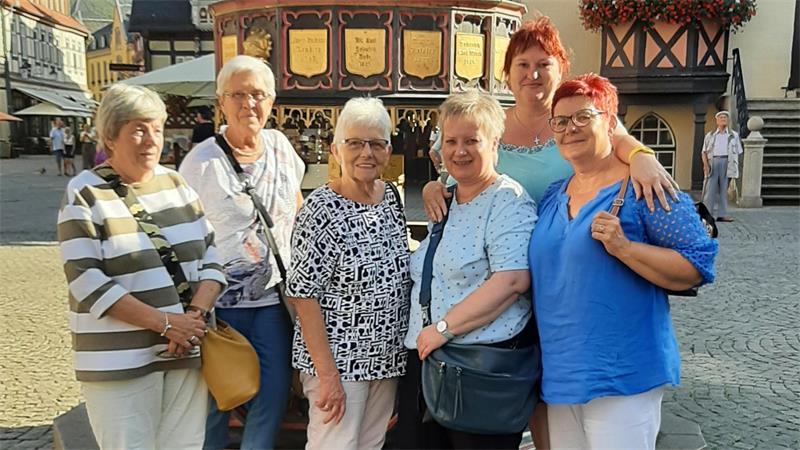 Sechs ältere Frauen stehen eng beieinander auf einem gepflasterten Platz vor Fachwerkhäusern und lächeln in die Kamera