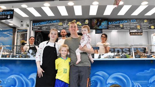 Familie mit zwei Kindern steht vor einem Fischbrötchen-Verkaufsstand mit blauem Wellendekor und beleuchtetem Deckenraster.