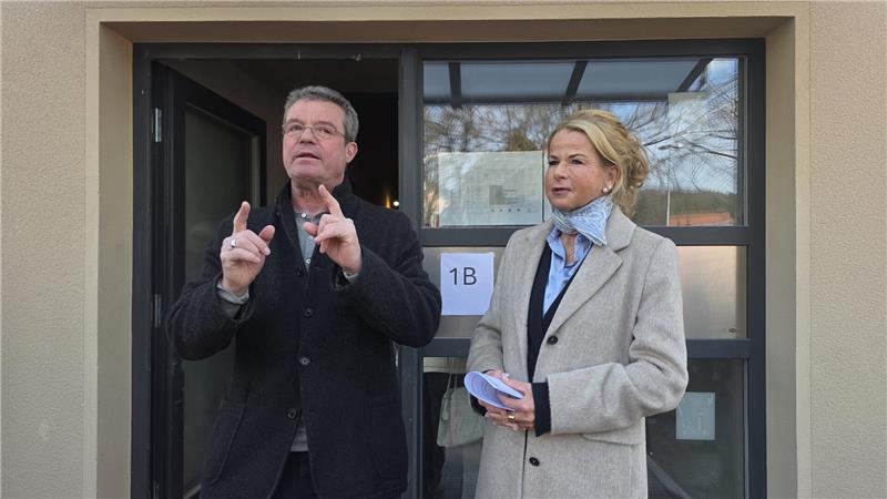 Zwei Personen stehen vor einem Gebäude mit der Beschilderung '1B', eine Person gestikuliert mit den Händen, die andere hält einen Stapel Karten oder Zettel.