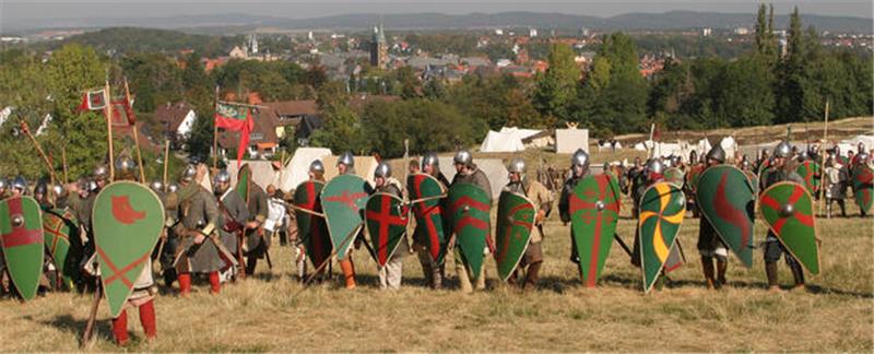 Gruppe von historisch gekleideten Rittern mit Schilden und Helmen auf einer Wiese vor einer Stadt im Hintergrund