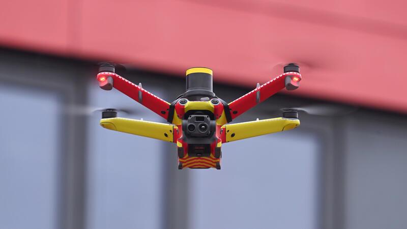 Nahaufnahme einer rot-gelb lackierten Drohne mit Kamera vor unscharfem Gebäudehintergrund.