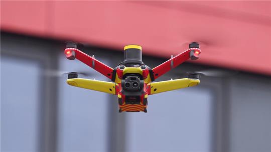 Nahaufnahme einer rot-gelb lackierten Drohne mit Kamera vor unscharfem Gebäudehintergrund.