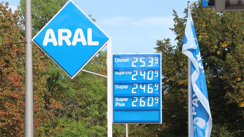 Tankstelle mit ARAL-Logo und Preistafel für Diesel, Super E10, Super und Super Plus vor Bäumen und blau-weißen Fahnen.