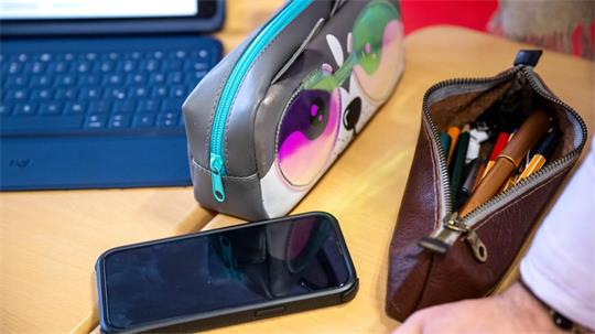 Auf einem Tisch liegen ein Smartphone, ein graues Federmäppchen mit Pandagesicht und ein geöffnetes braunes Federmäppchen mit Stiften