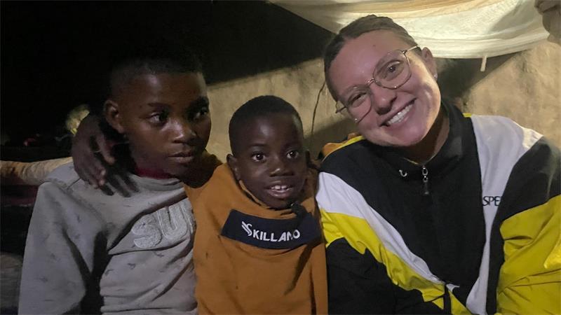 Drei Personen sitzen eng nebeneinander in einem dunklen Raum, eine Person trägt eine Jacke mit gelb-weißen und schwarzen Farbfeldern, eine andere ein orangefarbenes Shirt mit dem Schriftzug 'SKILLAND'
