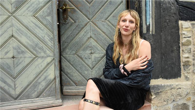 Eine Frau mit langen, blonden Haaren und schwarzem Kleid, das von einer Schulter rutscht, sitzt der Stufe vor einer Holztür.