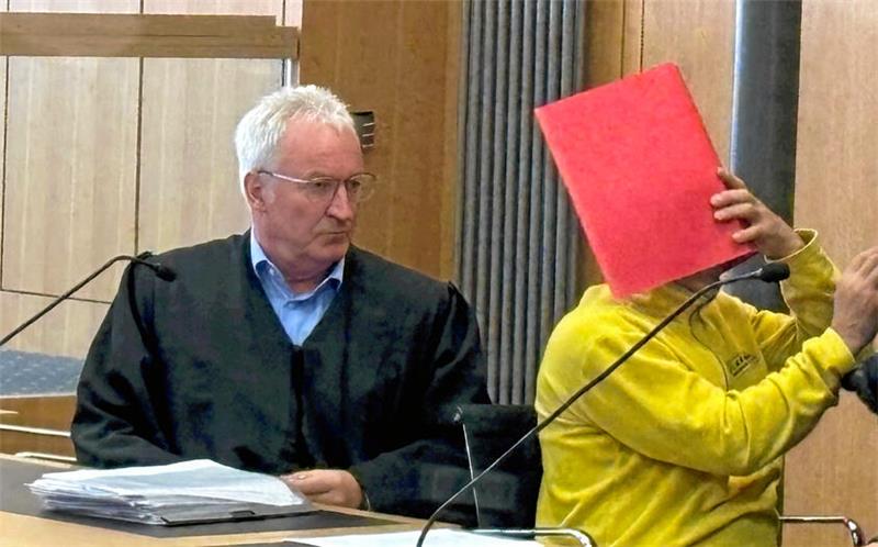 Zwei Männer sitzen an einem Tisch, einer trägt eine schwarze Robe, der andere eine gelbe Jacke und hält eine rote Mappe vor sein Gesicht.