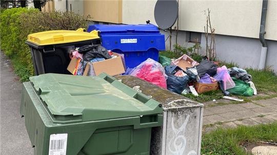 Mehrere Mülltonnen mit gelbem, blauem und grünem Deckel stehen vor einem Wohnhaus, daneben liegt eine große Menge Müllsäcke und Kartons auf dem Gehweg