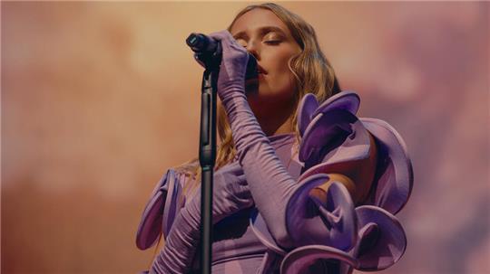 Sängerin mit langen Haaren und auffälligem, voluminösem Ärmelkleid singt vor Mikrofon