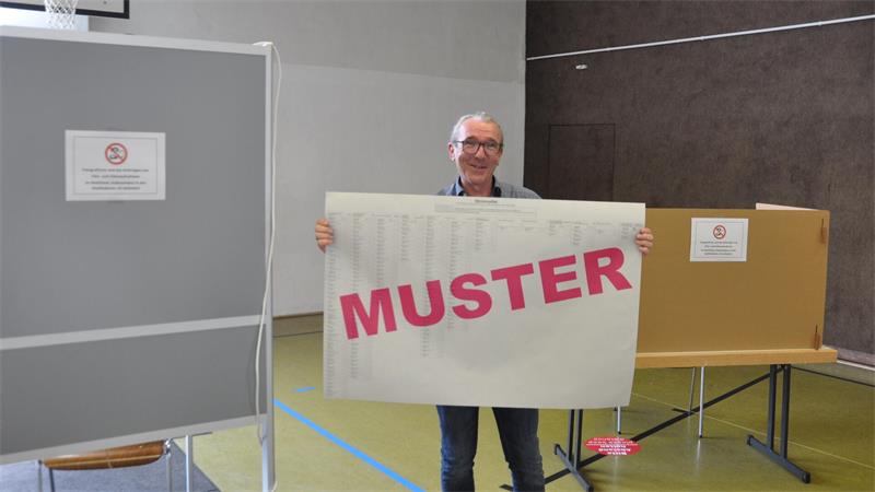 Älterer Mann steht in einem Raum und hält ein großes Schild mit rotem Schriftzug 'MUSTER'