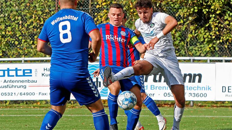 Drei Fußballspieler auf dem Spielfeld, zwei in blau-roten Trikots und einer in weißem Trikot, kämpfen um den Ball