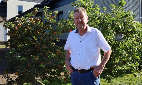 Mann in weißem Hemd und blauer Hose steht vor einem Apfelbaum in einem Garten vor einem Haus
