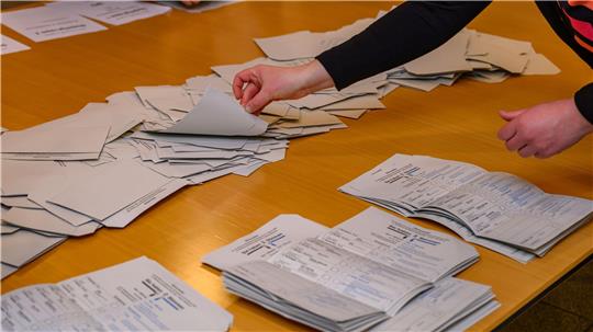 Das Foto zeigt die Hände einer Person, die weiter nicht auf dem Bild zu sehen ist, beim Auszählen von Stimmzetteln, die geordnet auf einem Tisch liegen.