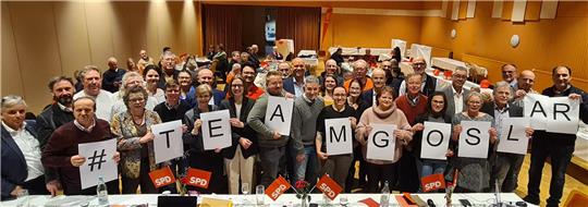 Gruppe von Menschen in einem Saal hält Schilder mit den Buchstaben '#TEAMGOSLAR' hoch