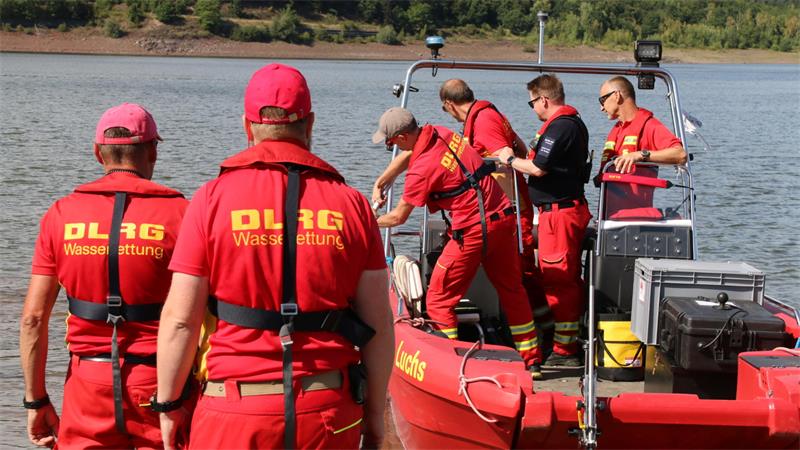 Mehrere Personen in roten DLRG-Wasserrettungsuniformen auf einem Boot und am Ufer eines Gewässers