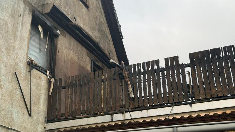 Haus mit brüchigem Holzbalkon und beschädigtem Fenster unter bewölktem Himmel.