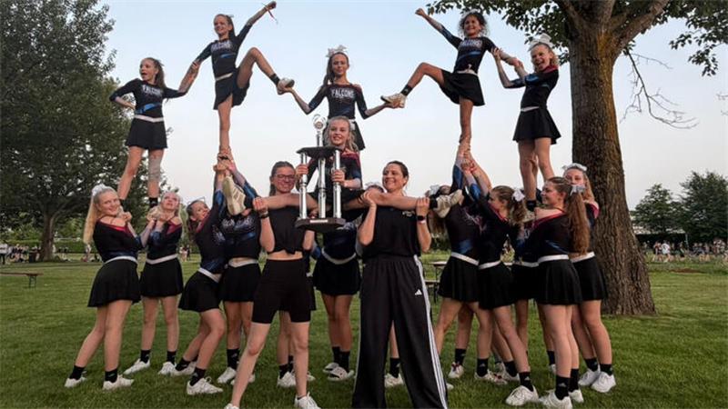 Gruppe von Cheerleaderinnen in schwarzen Uniformen posiert im Freien mit einer großen Trophäe und zeigt eine Pyramidenformation mit gehobenen Beinen