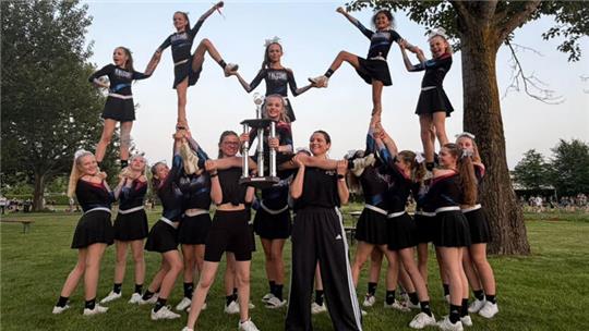 Gruppe von Cheerleaderinnen in schwarzen Uniformen posiert im Freien mit einer großen Trophäe und zeigt eine Pyramidenformation mit gehobenen Beinen