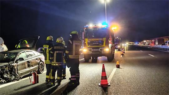 Feuerwehrmänner stehen neben dem beschädigten Unfallwagen und vor ihnen steht das Feuerwehrfahrzeug.