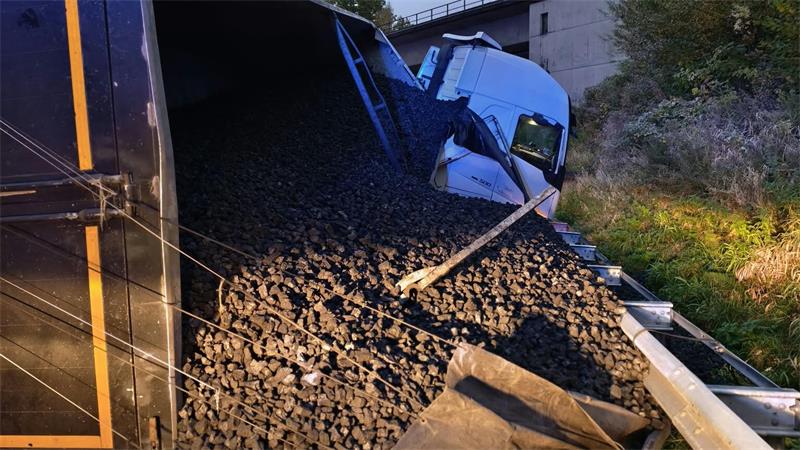 Umgekippter blauer Lkw auf der Seite mit ausgelaufenem Schotter neben einer Straße und Leitplanke bei bewölktem Himmel