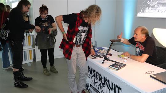 Person mit rotem Karohemd und schwarzem T-Shirt steht vor einem Tisch mit Büchern, an dem ein Mann mit schwarzem T-Shirt sitzt und mit der Hand gestikuliert, im Hintergrund stehen zwei weitere Personen in einer hellen Innenraumumgebung.