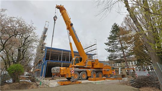Gelber Autokran auf einer Baustelle vor einem teilweise eingerüsteten Gebäude mit Bäumen im Hintergrund