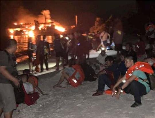 Nach der Rettung ruhen sich Überlebende an Land aus. Die Fähre brennt noch.
