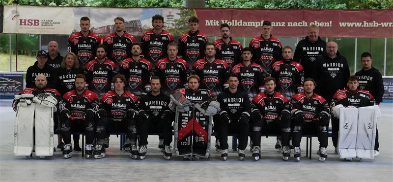 Gruppenfoto einer Eishockeymannschaft mit Spielern in schwarzen Trikots und zwei Torhütern in weißer Ausrüstung auf einer Eisfläche vor einer Anzeigetafel und Werbebannern