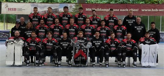 Gruppenfoto einer Eishockeymannschaft mit Spielern in schwarzen Trikots und zwei Torhütern in weißer Ausrüstung auf einer Eisfläche vor einer Anzeigetafel und Werbebannern
