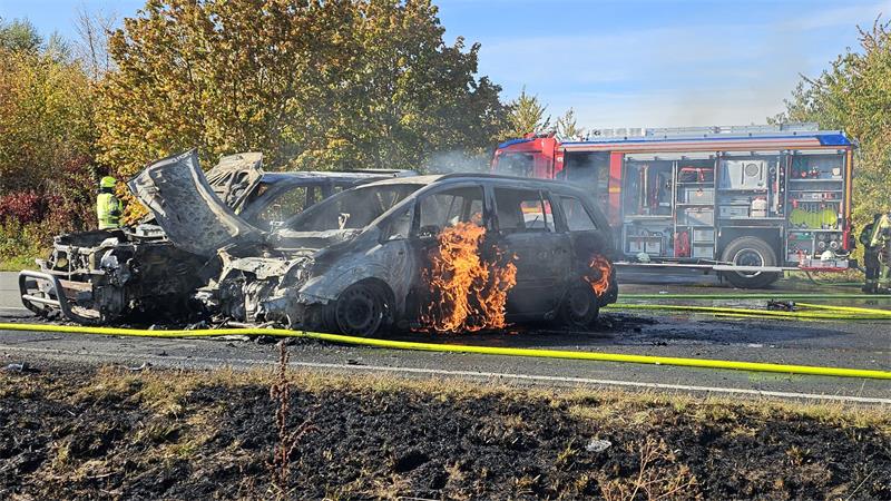 Brennendes Auto mit offener Motorhaube auf einer Straße, im Hintergrund Feuerwehrfahrzeug und Feuerwehrleute
