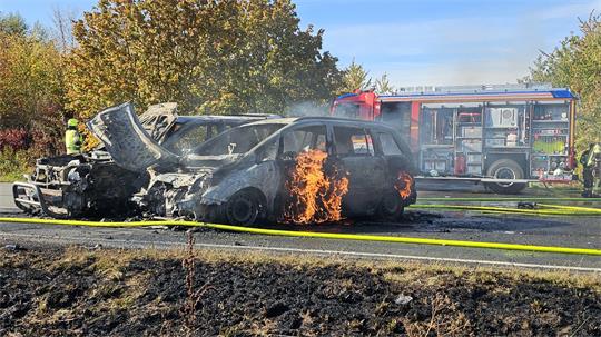 Brennendes Auto mit offener Motorhaube auf einer Straße, im Hintergrund Feuerwehrfahrzeug und Feuerwehrleute