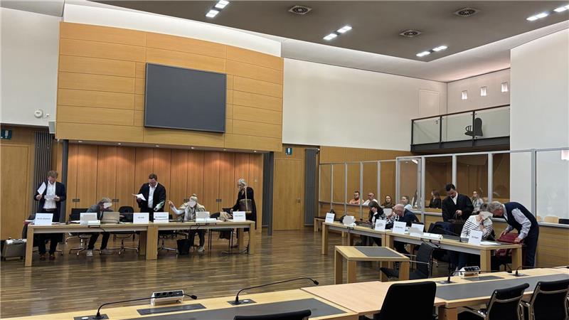 Großer Sitzungssaal mit mehreren Personen an Tischen, einige stehen, andere sitzen, im Hintergrund eine große Leinwand und Holzpaneele an der Wand.