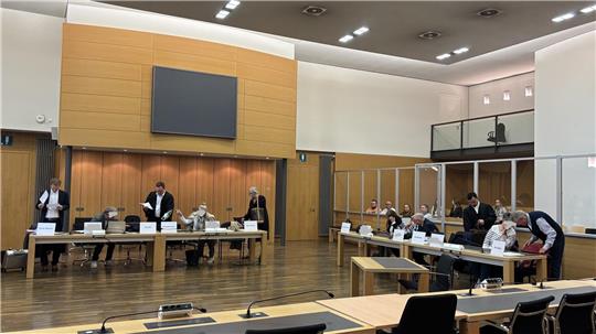 Großer Sitzungssaal mit mehreren Personen an Tischen, einige stehen, andere sitzen, im Hintergrund eine große Leinwand und Holzpaneele an der Wand.