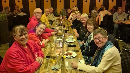 Gruppe von Menschen unterschiedlichen Alters sitzt an einem langen Holztisch in einem Restaurant und lächelt in die Kamera
